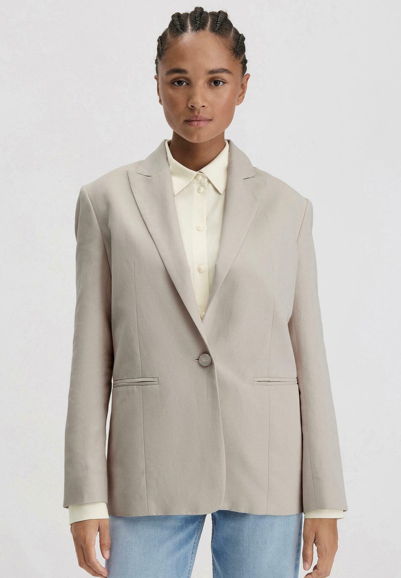 Beiger Blazer mit Reverskragen, Ein-Knopf-Verschluss und zwei frontalen Taschen. Getragen über einem hellen Hemd mit Knöpfen.