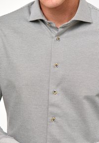 Mężczyzna w dopasowanej, jasnoszarej koszuli z małym, powtarzającym się wzorem geometrycznym i czterema widocznymi guzikami z przodu.