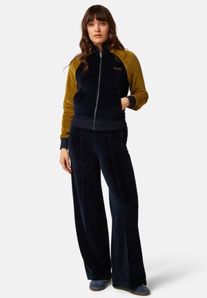 Mujer con el pelo largo y castaño que lleva una chaqueta con cremallera con mangas mostaza y pantalones azul marino, de pie con una mano en el bolsillo.