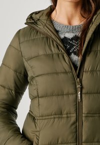 Olīvu zaļš pūķa jaka ar rāvējslēdzēju priekšā, ar kapuci un quilted tekstūru. Uzvilkts virs pelēka rakstainā džempera.