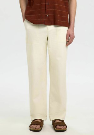 Persoon die crèmekleurige rechte broek draagt met grote voorzakken, bruine sandalen met open tenen en een roestkleurig overhemd met knopen.