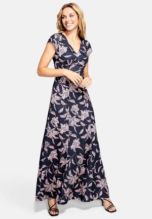 Smilende kvinde iført en marineblå maxikjole med pink blomsterprint og korte ærmer, kombineret med sorte remsandaler med hæl.