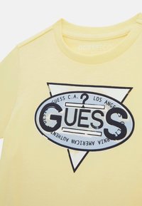 Sárga organikus pamut póló, nagyméretű, ovális logóval, amelyen a "GUESS" felirat és fekete, kék grafikai részletek találhatók, valamint háromszög mintázat.