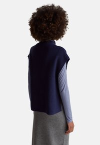 Osoba s kudrnatými vlasy, oblečená v námořnicky modrém svetru bez rukávů přes dlouhý modrý rukáv a šedou sukní, stojící zády k jednoduchému pozadí.