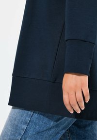 Felpa blu navy con bordi e orlo a coste, texture del tessuto liscia e design casual, parzialmente mostrata con una mano appoggiata sul fianco.