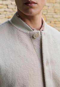 Light gray textured jacket with a soft, fuzzy surface, featuring a ribbed collar, two buttons, and a visible patterned inner layer.
