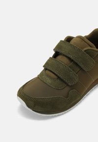 Zelena tenisica koja se sastoji od kombinacije glatkih i teksturiranih materijala, s dva podesiva Velcro čepa i bijelom gumenom potplatom.
