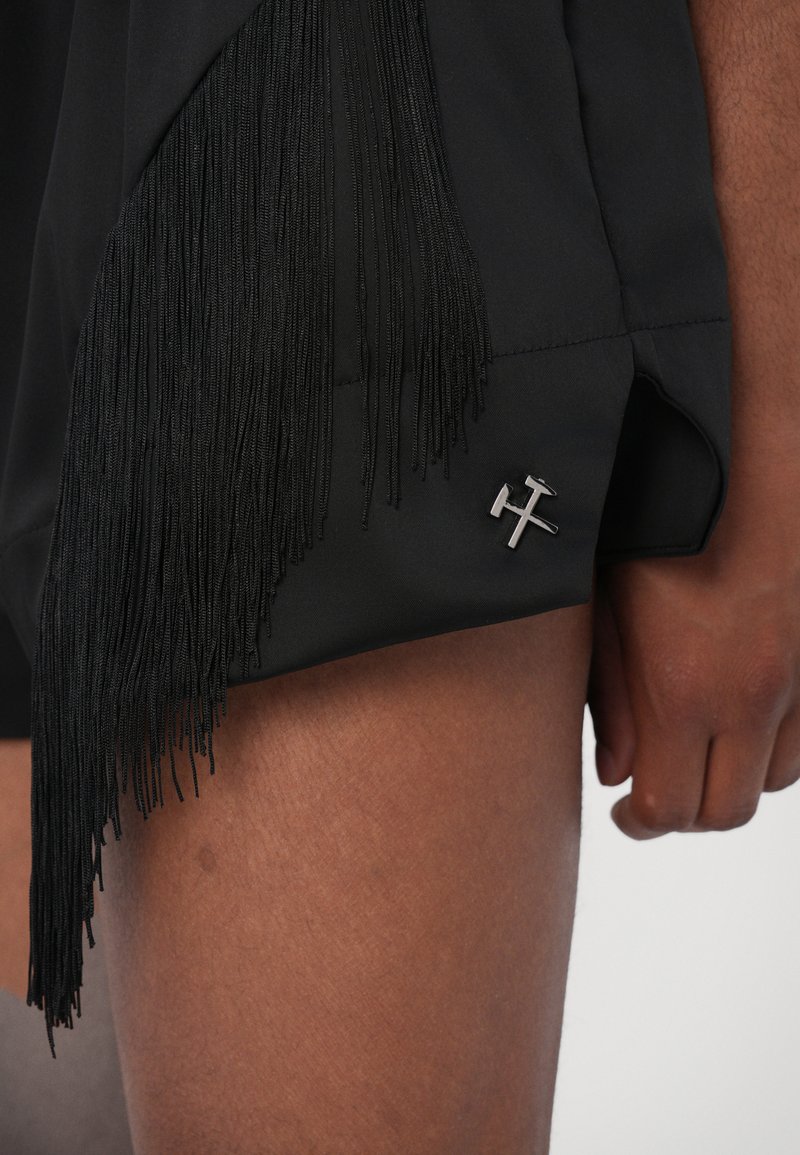 Sorte shorts med lange sorte frynser langs kanten, med en sølvfarvet krydsformet udsmykning på siden. Glat stoftexture.