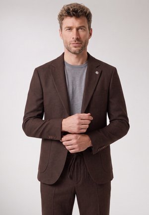 Man met kort krullend haar die een bruin colbert en broek draagt over een grijs overhemd, zijn mouwen verstelend, staand tegen een eenvoudige achtergrond.