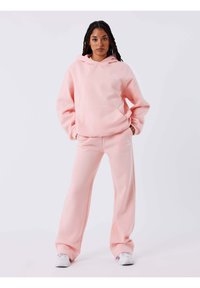 Lichtroze hoodie met een capuchon, een zak aan de voorkant en een wit logo, gecombineerd met bijpassende losse broek. Heeft een zachte, gebreide textuur.