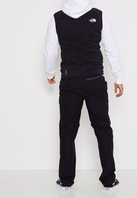 Eine Person trägt eine schwarze Weste von The North Face über einem weißen Kapuzenpullover, schwarze wandelbare Hosen und schwarze Sneakers mit niedrigem Schnitt und steht mit dem Rücken zur Kamera.