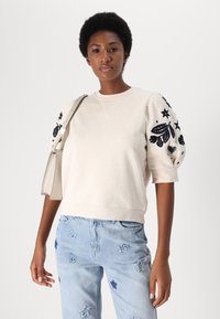 Lichtbeige sweatshirt met pofmouwen, voorzien van marineblauwe bloemen- en sterrenborduurwerk. Gecombineerd met lichtblauwe jeans versierd met steraccenten.