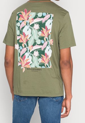 Henkilö, joka käyttää oliivinvihreää T-paitaa, jossa on suuri värikäs trooppinen kukkakuosi ja takana teksti "Scotch & Soda Amsterdam".