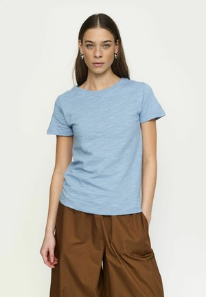 Žena s dlhými hnedými vlasmi má na sebe svetlomodrú krátku košeľu s rukávmi a voľné hnedé nohavice, stojaca pred jednoduchým bielym pozadím.