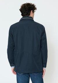 Man with dark curly hair wearing a navy blue jacket and blue jeans, standing facing away against a plain white background.