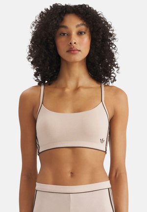 Vrouw met krullend haar, gekleed in een beige cropped top en bijpassende broek met bruine bies, staand tegen een effen witte achtergrond.