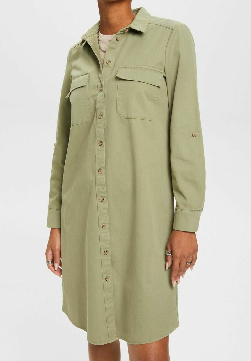 Femme portant une robe chemise vert olive boutonnée, à manches longues, longueur genou, avec deux poches poitrine et un col.