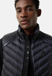 Czarna pikowana kamizelka z przeszyciami w kształcie litery V, wysokim kołnierzem i zamkiem błyskawicznym z ozdobnym suwakiem. Gładka faktura z odblaskowymi akcentami.