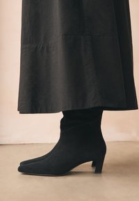 Schwarze Veloursleder-Stiefeletten mit spitzer Zehenpartie und niedrigem Blockabsatz, kombiniert mit einem langen schwarzen Rock. Die Absatzhöhe ist mittelhoch.