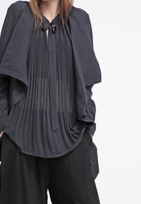 Czarny warstwowy top z plisowaną teksturą, wiązaniem przy szyi i przezroczystymi panelami, uzupełniony szerokimi czarnymi spodniami. Miękka tkanina, luźny krój.