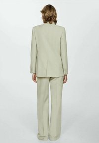 Persoon met schouderlang haar in een lichtbeige pak, staande met de rug naar de camera tegen een effen witte achtergrond.