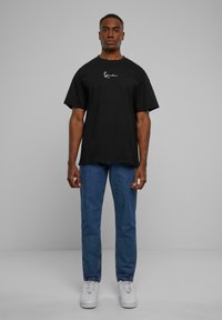 Black short-sleeve t-shirt with a white graphic design on the chest, paired with blue jeans and white sneakers. Plain background.