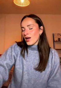 Młoda kobieta z długimi brązowymi włosami i srebrnymi kolczykami w kształcie kół, ubrana w szary sweter, mówi lub śpiewa w pomieszczeniu przy ciepłym świetle.