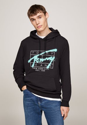 Felpa nera con tasca frontale, caratterizzata da un logo grafico turchese e testo. Indossata sopra una maglietta bianca, abbinata a jeans blu.