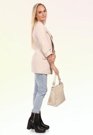 Femme aux longs cheveux blonds portant un blazer beige, un jean clair, des bottines noires à talons, tenant un sac à main beige sur fond blanc.