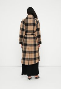 Manteau long à carreaux en mélanges de laine beige, noir et gris. Doté d'un col, d'une taille ceinturée et d'une fente dans le dos. Porté sur une robe noire.