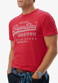 Rode katoenen t-shirt met een ronde hals, voorzien van witte tekst en graphics. In combinatie met een geruit overhemd dat om de taille is geknoopt en een bruine riem.