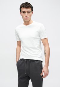 Fehér pamut rövid ujjú póló, kerek nyakkivágással, sötétszürke, gumis derékrészű és húzózsinórral ellátott melegítőnadrággal viselve.