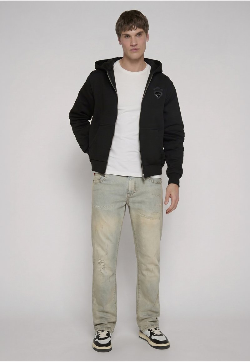 Jeune homme portant un sweat à capuche zippé noir sur un t-shirt blanc, un jean clair usé et des baskets noires et blanches, debout devant un fond uni.