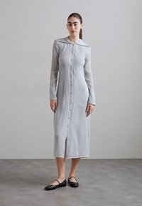 Vestido cinza claro, com botões, gola, mangas longas, tecido texturizado e fenda lateral. Usado com sapatos planos pretos sobre um fundo neutro.