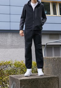 Svart zip-up hoodie med framficka och matchande svarta byxor, båda i mjukt tyg. Bärs med vita sneakers, stående på en betongplatta.