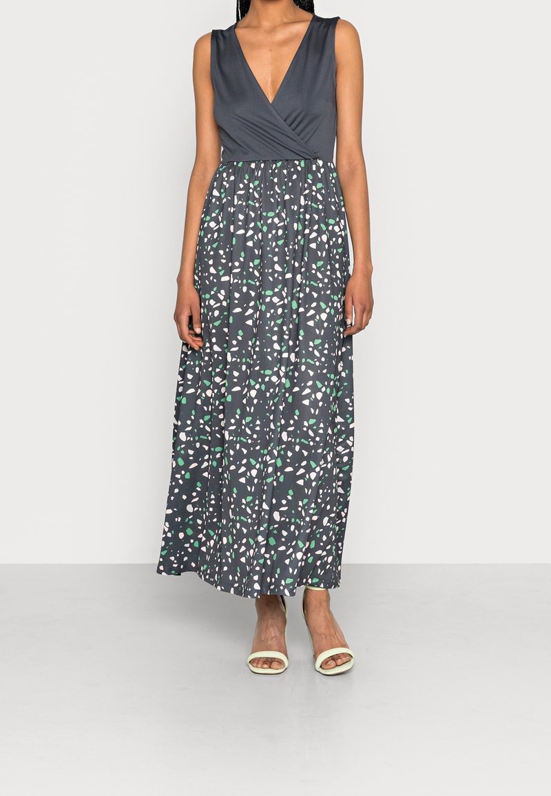 Femme portant une robe maxi sans manches à col en V avec un haut uni gris foncé et une jupe à motifs blancs et verts, associée à des sandales claires.