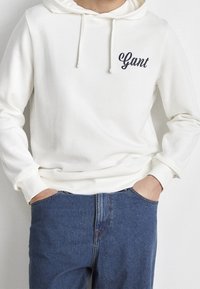 Bílá mikina s kapucí a šňůrkami, s tmavě modrým logem "Gant". V kombinaci s modrými džínami, které ukazují ležérní střih a texturu.