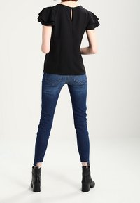 Black blouse with ruffled sleeves and back keyhole detail, paired with fitted dark blue jeans and black ankle boots, showcasing contrast textures.