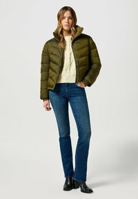 Giacca a vento verde con zip frontale, caratterizzata da un collo alto e un design a righe orizzontali. Abbinata a un maglione crema e jeans blu.