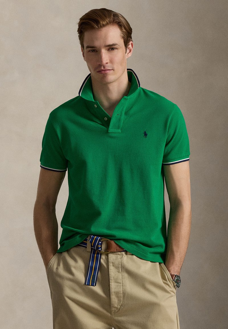Πράσινο πόλο πουκάμισο από υλικό μ ε υφή, με γιακά δύο κουμπιών, αντίθετο μπλε σκούρο περιθώριο και μικρό λογότυπο. Συνδυασμένο με μπεζ παντελόνι και ριγέ ζώνη.