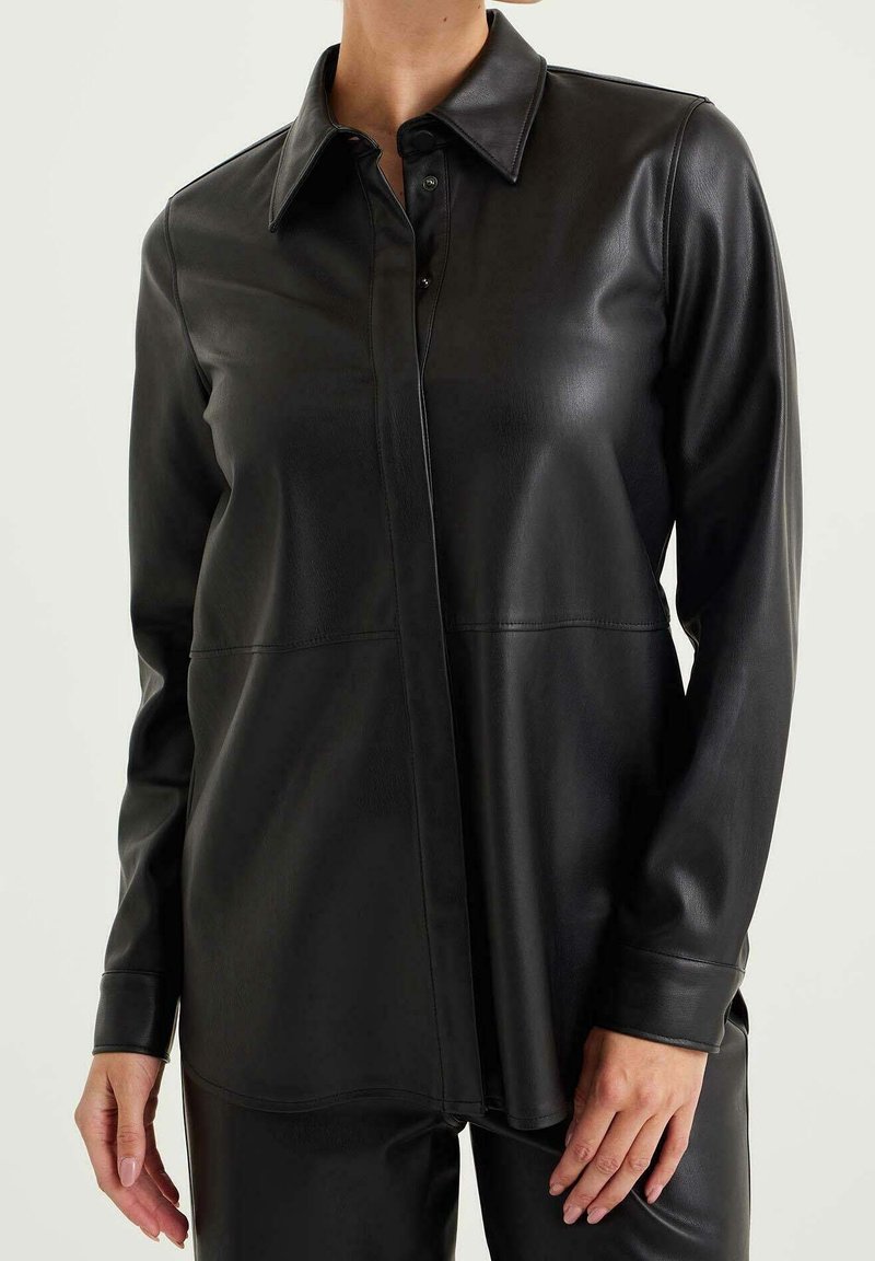 Kvinna klädd i en långärmad svart skjorta i konstläder med knappar och matchande byxor, stående framför en enkel ljus bakgrund.