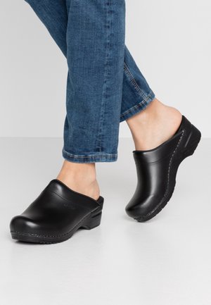 Personne portant des sabots noirs en cuir à talons en bois et un jean bleu, debout sur un sol blanc devant un fond uni.