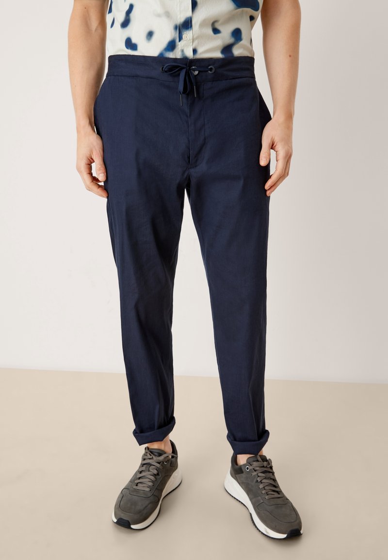 Mörkblå linnebyxor med elastisk midja, smalnande ben och upprullade fållar, i kombination med grå träningsskor.