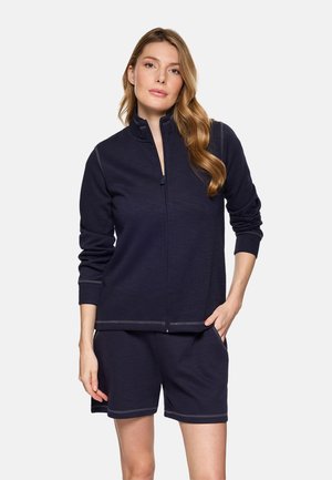 Mujer con cabello largo y ondulado, vestida con una chaqueta azul marino con cremallera y pantalones cortos a juego, de pie con una mano en el bolsillo contra un fondo liso.