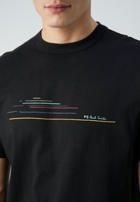 Gros plan sur un T-shirt noir avec des lignes horizontales colorées et le texte "PS Paul Smith" sur la poitrine, porté par une personne.