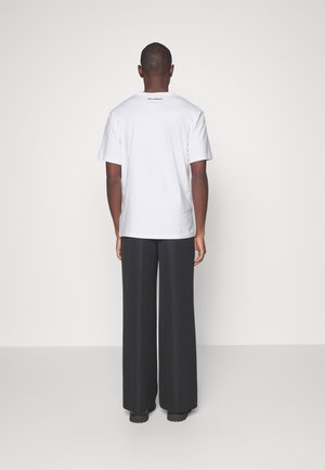 Camiseta de algodón blanca con mangas cortas y cuello redondo. La parte posterior presenta un pequeño detalle de logo negro. Combinada con pantalones negros de longitud completa.