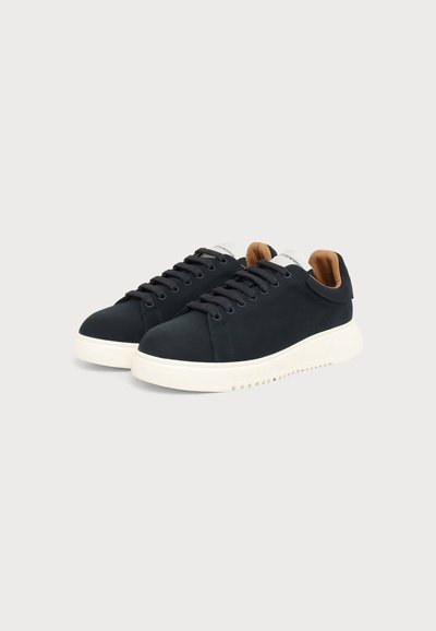 Paire de baskets basses noires avec semelles épaisses blanches et doublure intérieure beige sur un fond clair uni.