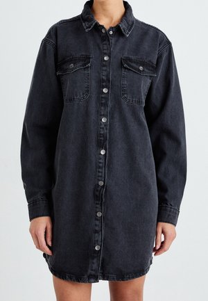 Robe-chemise en denim noir à manches longues, avec fermeture à boutons sur le devant et deux poches à rabat sur la poitrine, portée par une personne sur un fond uni.