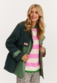 Zelená a námořnická modrá voděodolná bunda s páskem v pase, růžovým prošíváním a štítkem; vrstvená přes růžový pruhovaný svetr a kalhoty s leopardím vzorem.