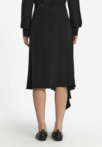 Svart midi kjol med asymmetrisk nederkant, slät textur och sidofäste detalj. Bärs med svarta platta skor.